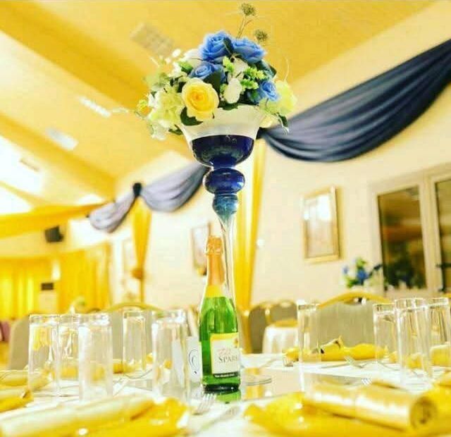 Tall glass vase centrepiece with blue roses and yellow flowers on a hall table with yellow napkins, with navy and gold drapes behind