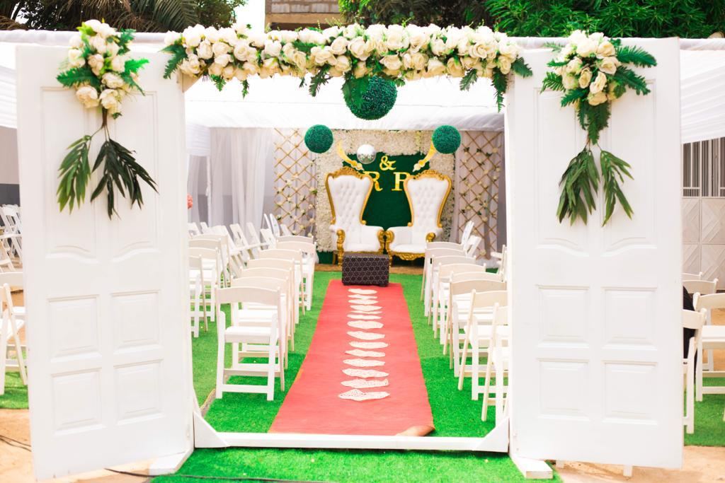 Two white doors dressed with white roses and green palm leaves forming an entrance, with a red aisle, white chairs and gold thrones in the distance