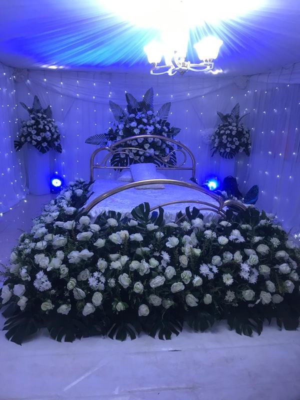White roses and green monstera leaves arranged around a bed frame, with fairy-light curtains and blue lighting, styled as a memorial tribute