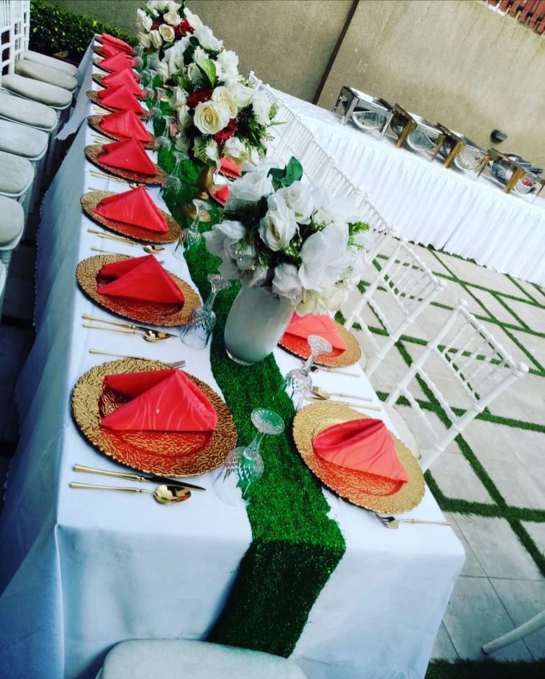 Long white-clothed table with a green runner, gold chargers, coral folded napkins and vases of white and red roses beside white chairs