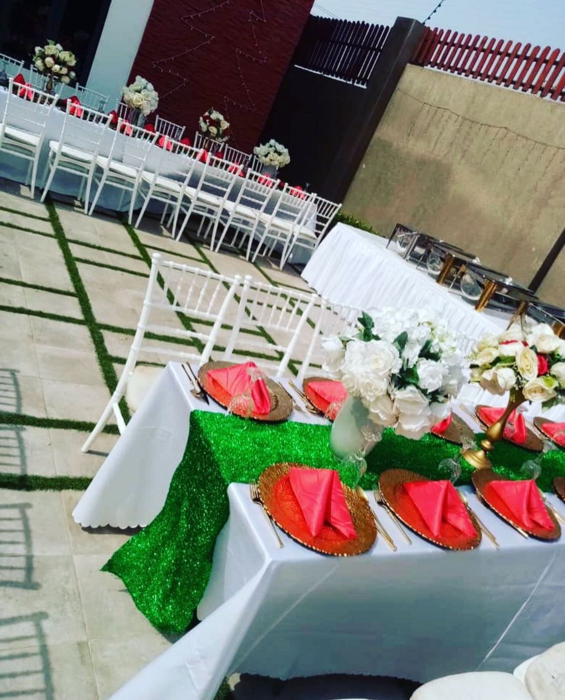 Outdoor banquet tables with white linen, green glitter runners, coral napkins on gold chargers and white flowers, with white chairs in rows