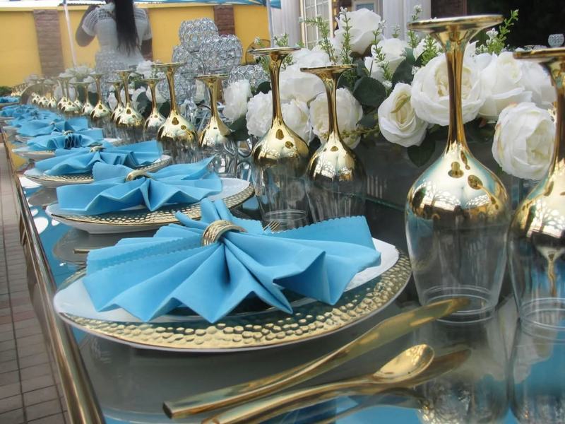Close view of a long glass-topped table set with gold hammered chargers, folded sky-blue napkins in gold rings, upturned gold goblets and white roses