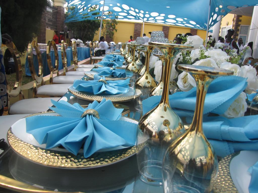 Teal and gold table setting with white roses and folded blue napkins under a blue canopy, styled by Flower Palace Decor Ghana