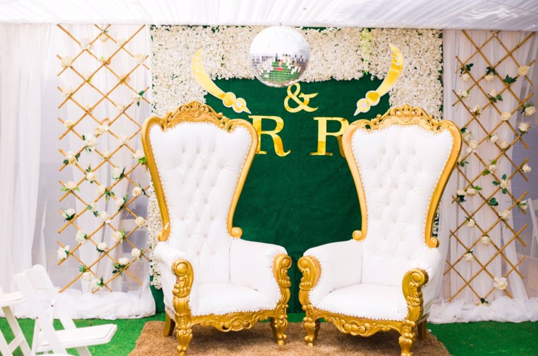 Two gold-framed white throne chairs in front of a white flower wall and green backdrop with gold lettering, set up for a ceremony