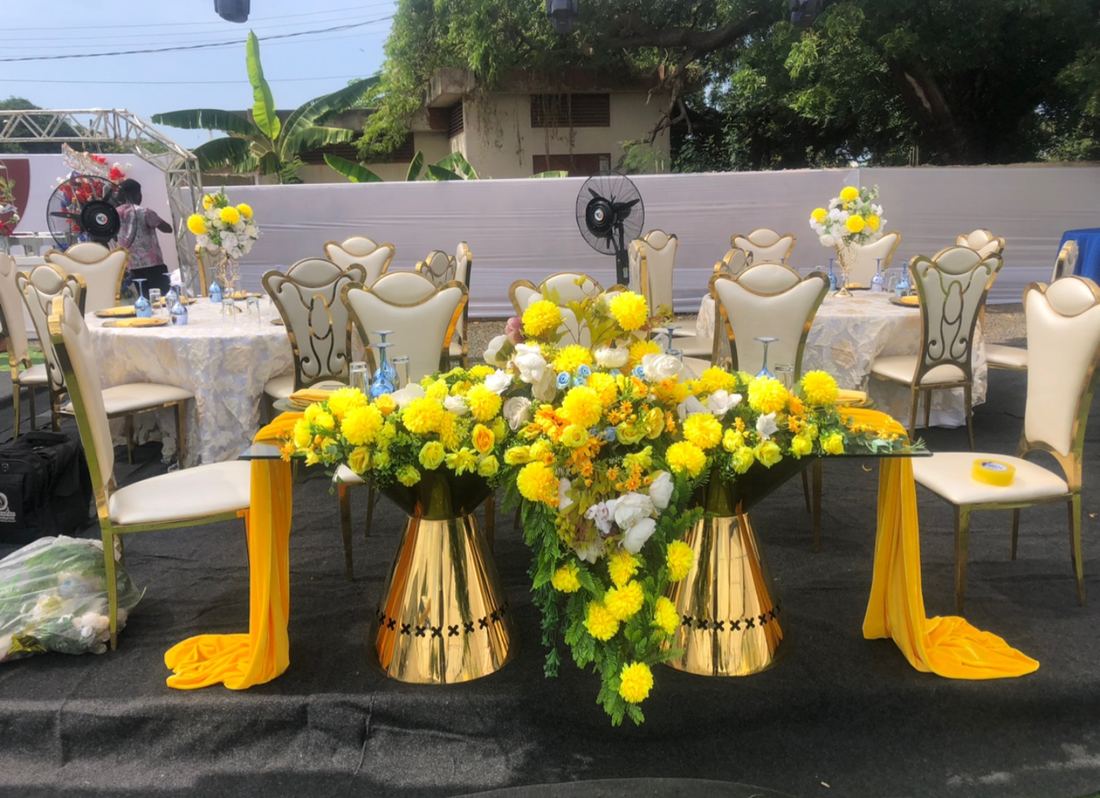 Glass-topped table on two gold pedestals with a large arrangement of yellow and white flowers, yellow drapes and cream gold-framed chairs