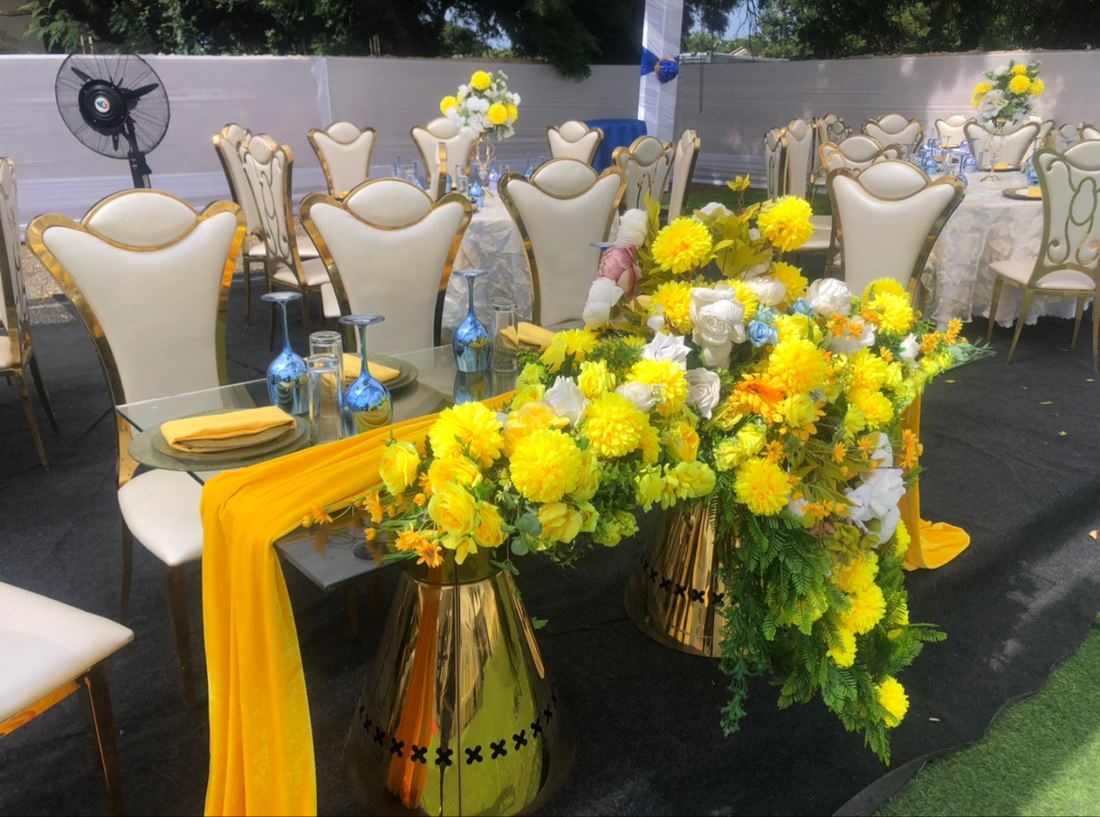 Side view of a glass table on gold pedestals with yellow flowers and green foliage, cream gold-framed chairs and round tables behind